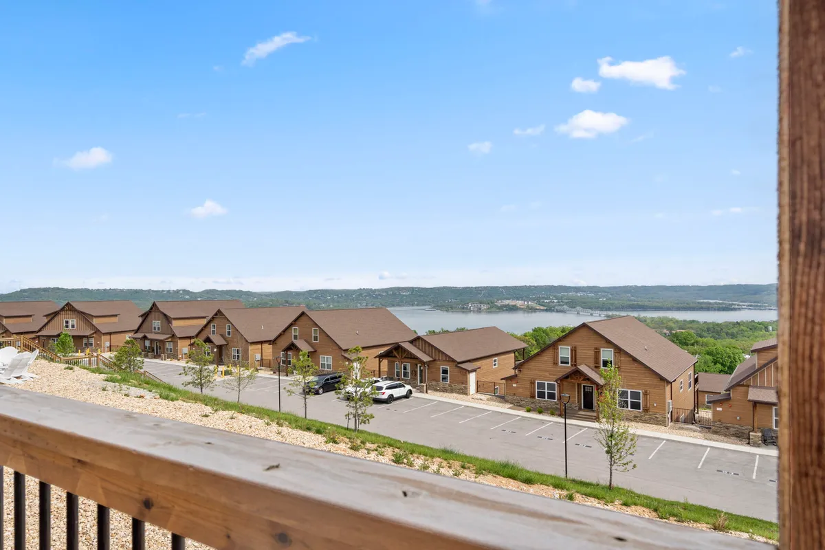 A serene exterior view capturing a row of houses with a tree-lined backdrop, parking area, and a peaceful lake beyond. The wide, open lawn and walking paths suggest a space ideal for strolls, outdoor activities, and community gatherings.