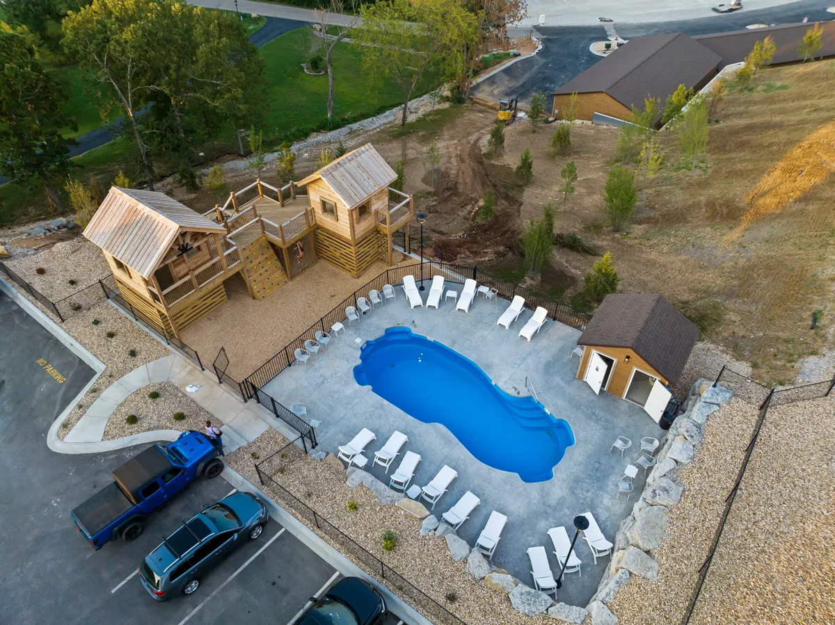 This image captures a vibrant outdoor pool area surrounded by lounge chairs and lush greenery, with visible playground structures nearby. The scene is filled with natural light and suggests the bustle of families and children enjoying the water and the play area.