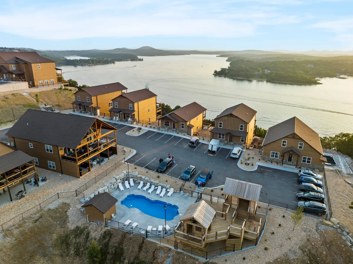 This image features a vibrant outdoor area with a swimming pool, a playground structure perfect for children, a clear view of the lake, and cozy wooden cabins, all under an open sky. The scene captures families swimming, children playing, and groups relaxing by the water, surrounded by nature.