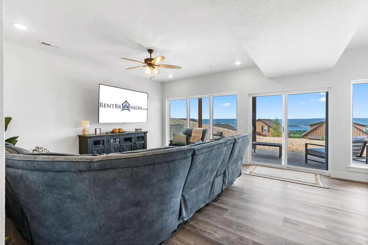 A spacious downstairs living room featuring a large sectional sofa, flat-screen TV, panoramic windows, and a decorative cabinet. The space is designed for comfort, with ample seating for groups to gather for movie marathons or game nights.