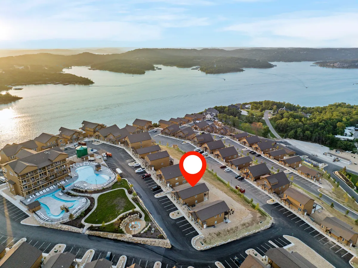 A wide-angle exterior shot capturing the swimming pool with a sweeping lake view in the background, bordered by a row of modern houses and a spacious parking area. The scene conveys both natural beauty and easy access to water adventures.