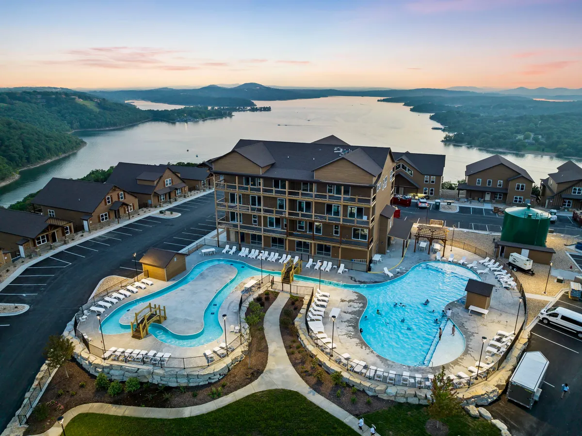 This image features multiple swimming pools with lounge chairs and landscaping, with a view of the lake and building facades in the background. The setting is outdoors and highlights both aquatic recreation and natural beauty. People can be seen socializing and relaxing poolside as the sun sets over the water.