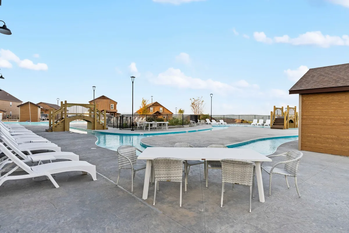 The scene showcases a lively outdoor setting with a swimming pool, an outdoor dining table set for group meals, a wooden bridge for playful exploration, and a playground structure. Families and children are visible enjoying both structured and spontaneous activities in a lush, inviting environment.