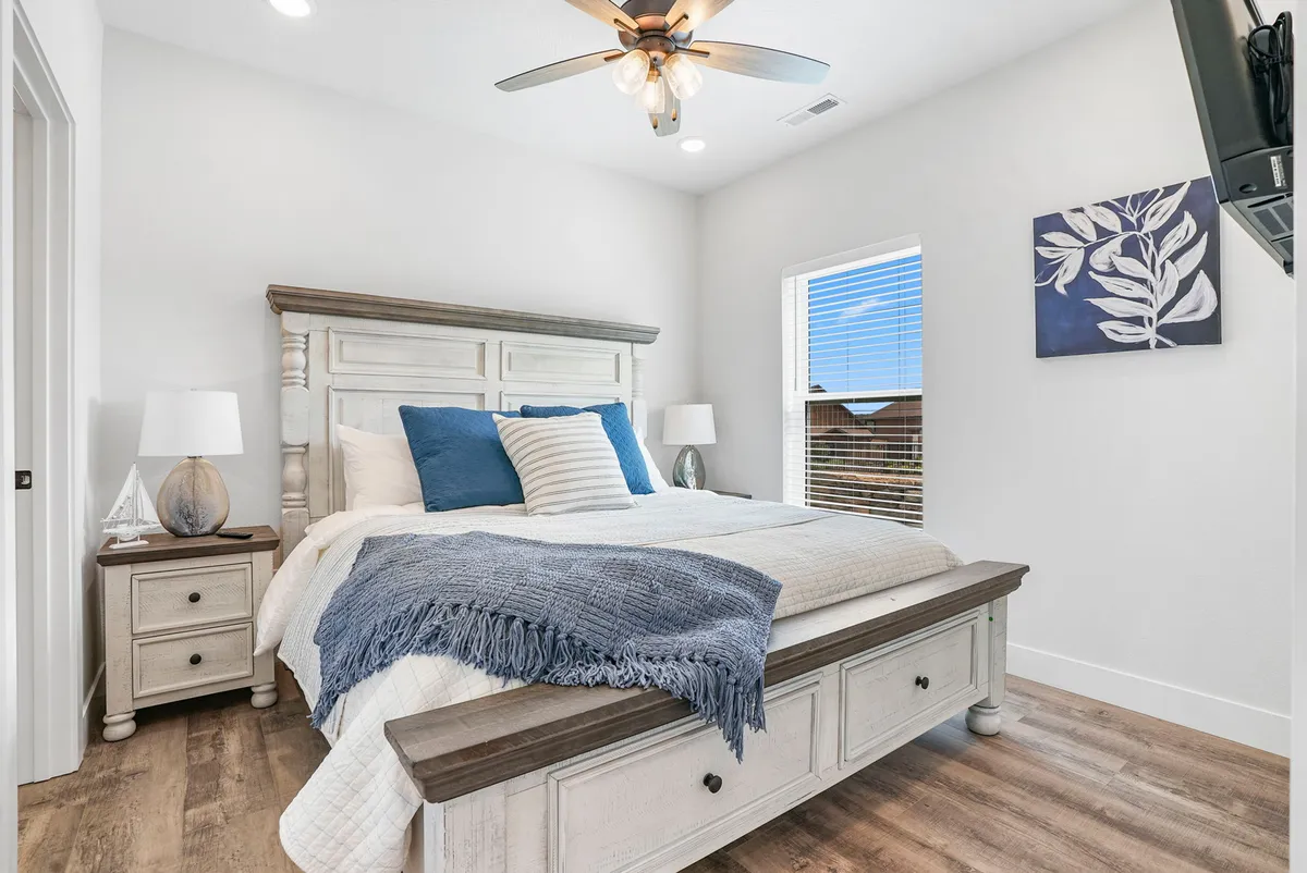 This image presents a cozy bedroom featuring a bed with decorative pillows, a nightstand with a lamp, and artwork on the wall. Sunlight filters through a window, creating a warm and serene ambiance ideal for relaxation.
