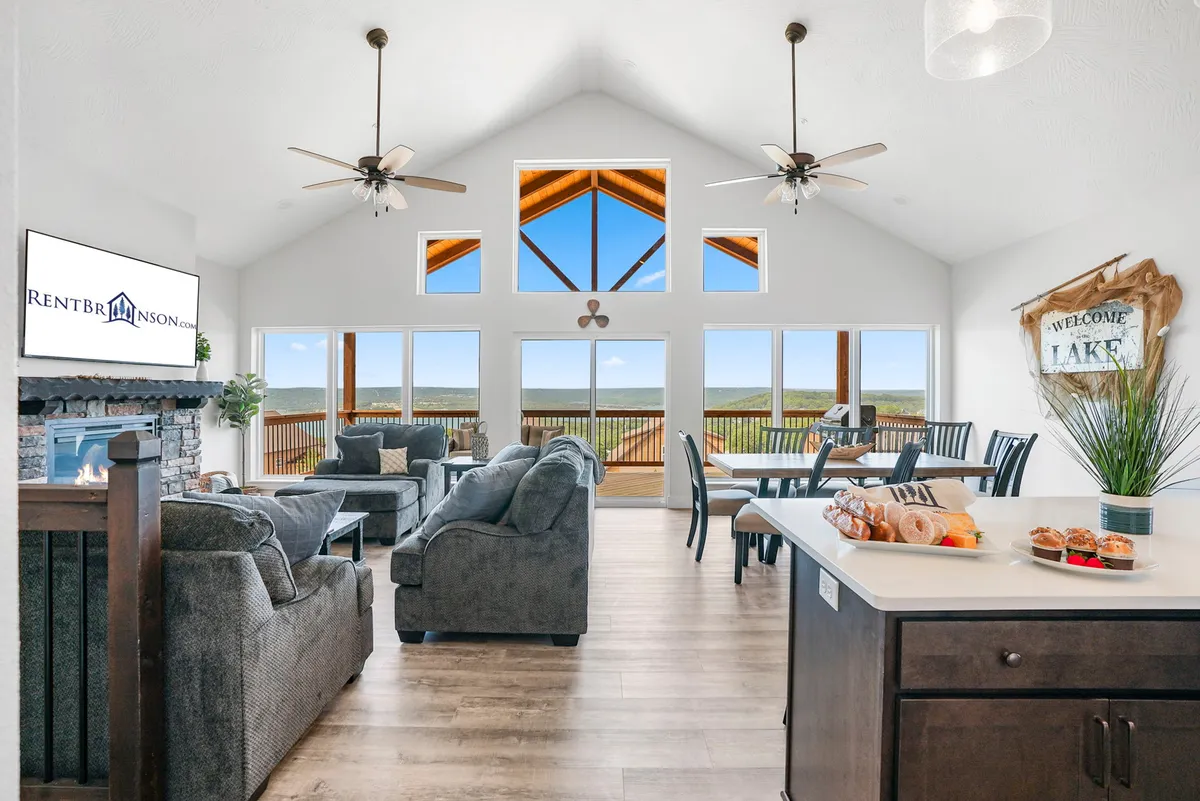 This inviting interior showcases spacious, modern furnishings, a welcoming dining area, large windows framing breathtaking lake views, a cozy fireplace, and a kitchen counter set with snacks—perfect for family gatherings and relaxation.